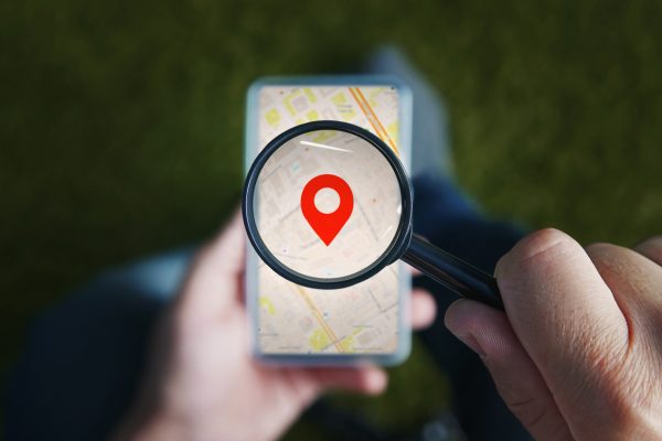 Shot of male hands holding magnifying glass with the red icon of geo-location and looking through it to the smartphone with online city map on the screen. Concept of store locator app and finding a business with search engine.